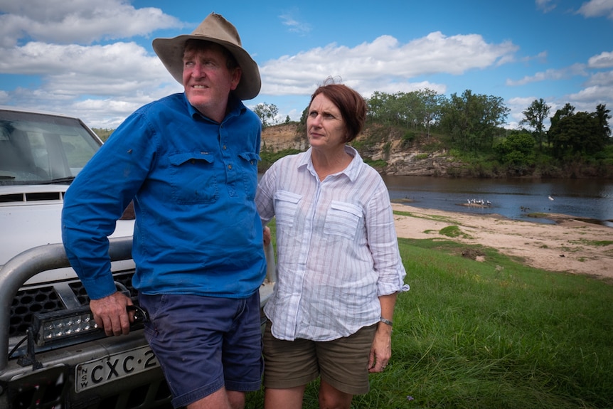 A man wearing an old hat and a woman lean against a ute parked on their property with a river scene in the background.