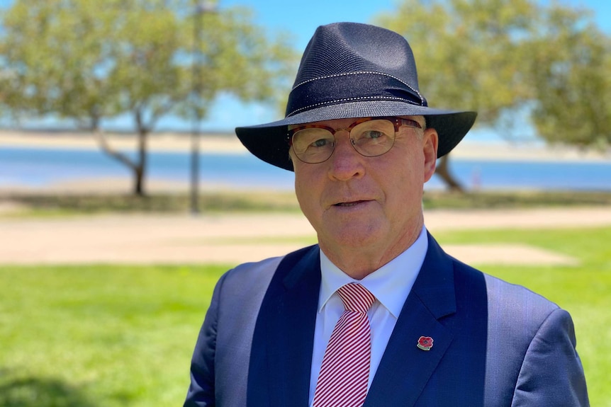 An older man dressed in a suit and wearing a hat, standing outside near a waterway.