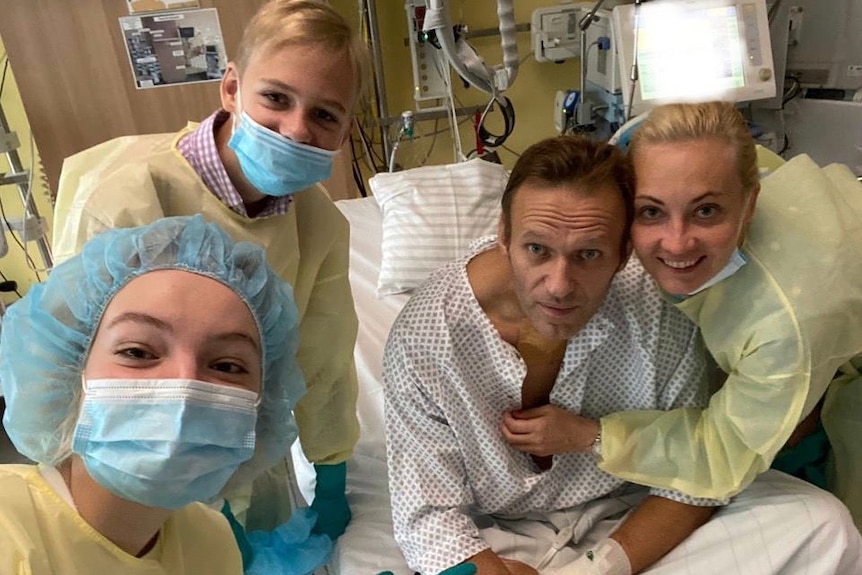 Alexei Navalny sits in a hospital bed in a medical gown surrounded by three people in protective gowns, masks and hair nets