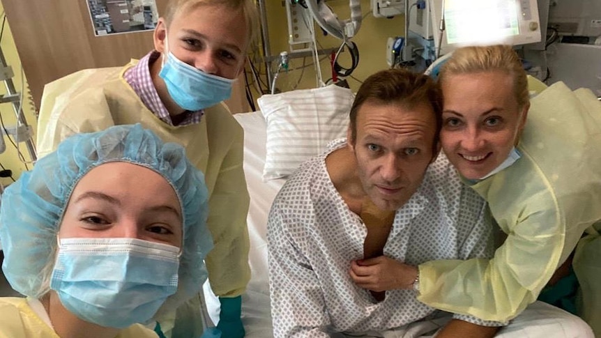 Alexei Navalny sits in a hospital bed in a medical gown surrounded by three people in protective gowns, masks and hair nets