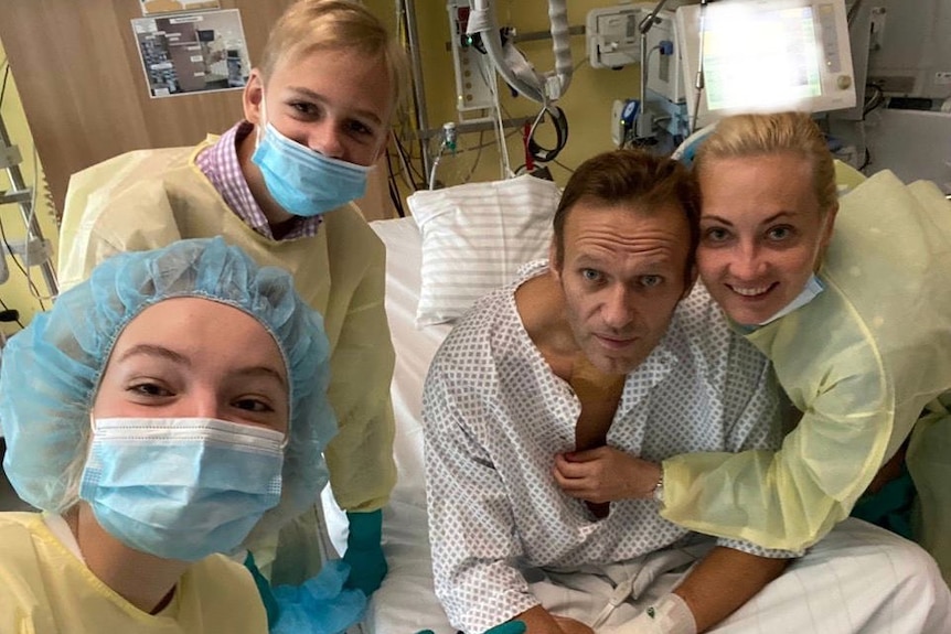 Alexei Navalny in einem Krankenhausbett in einem medizinischen Kleid, umgeben von drei Personen, die Schutzkleider, Masken und Haarnetze tragen.