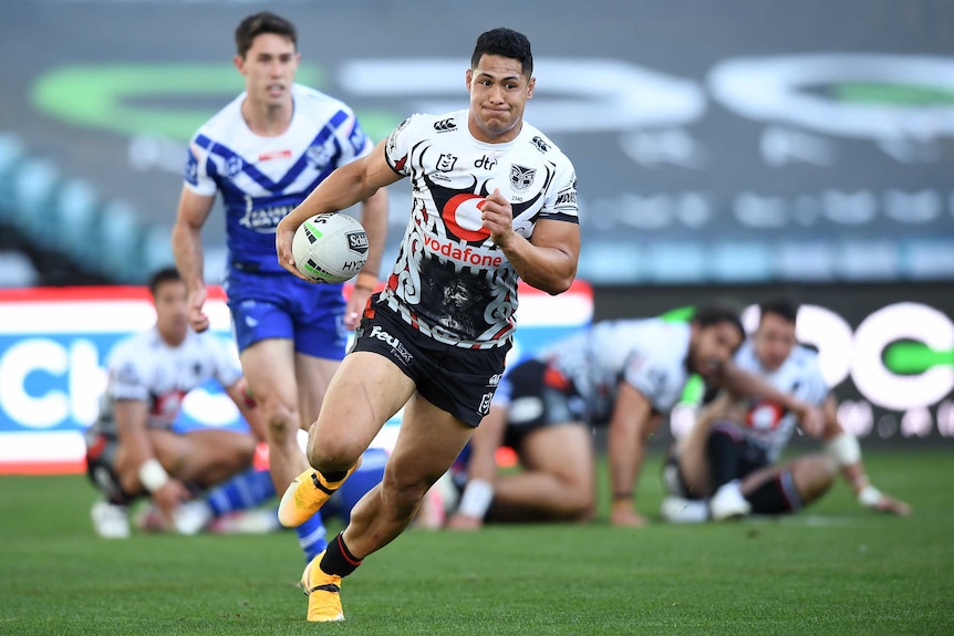 A Warriors NRL player runs with the ball; in his right hand as a Canterbury opponent gives chase in the background.
