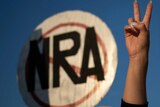 A hand gives a peace sign in front of a round sign with the letters NRA crossed out.