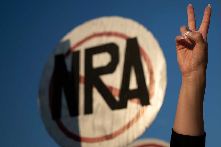 A hand gives a peace sign in front of a round sign with the letters NRA crossed out.