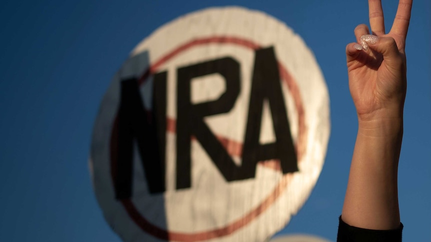 A hand gives a peace sign in front of a round sign with the letters NRA crossed out.