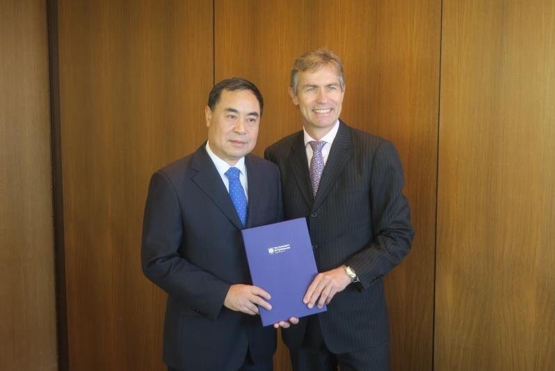 Two men, wearing black suits, pose for a formal photo. They are holding a document between them.