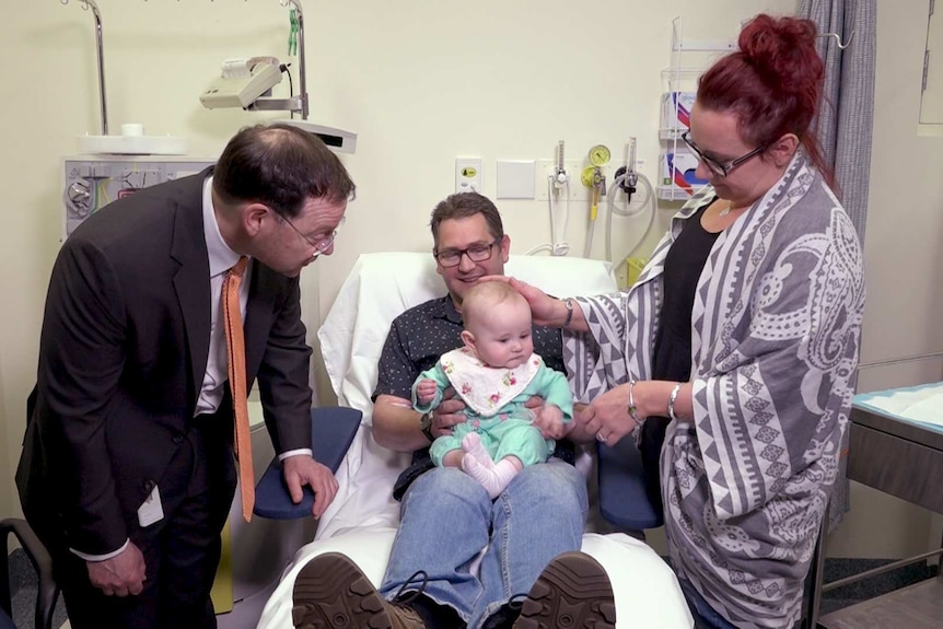 Porfessor John Rasko looks at baby Violet sitting on the knee of Mark Lee as he lays in a hospital bed next to partner Shannon.