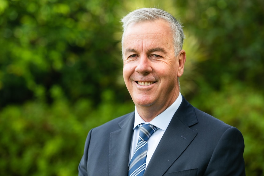 A man in a suit and white tie smiling at the camera with green bushes in the background.