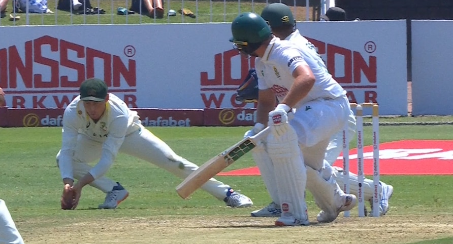 A cricketer takes a low catch with the ball hitting grass through  his fingers.