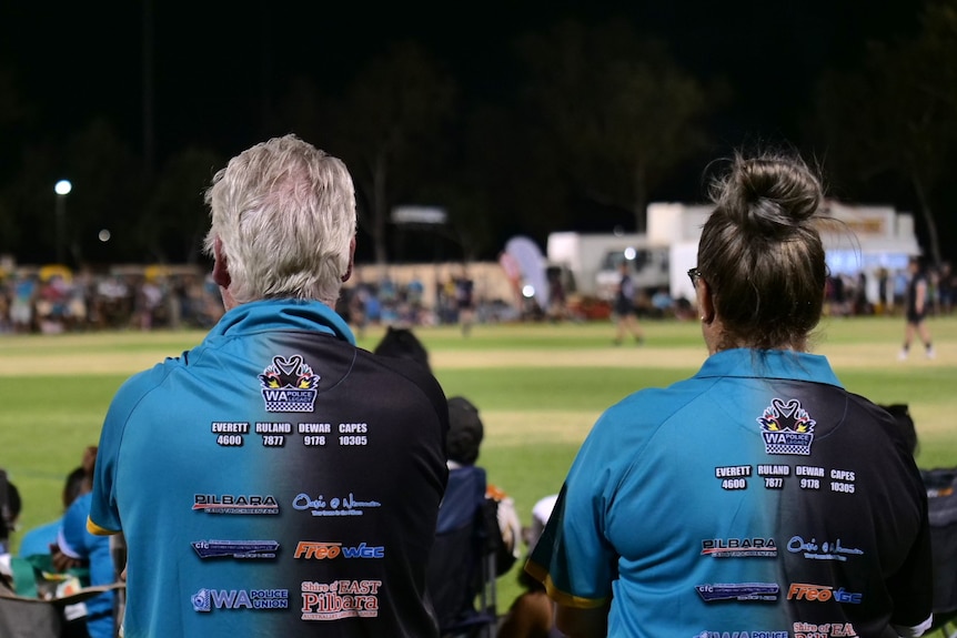 Two people from behind wearing light blue sports tops while watching a rugby game.