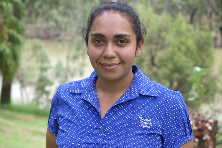 Dani Beezley, smiles at the camera, wearing blue shirt with white dots, trees and river behind.