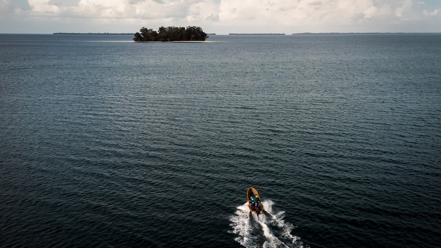 An aerial view shows a motor boat heading out towards an island in the middle of a calm blue sea