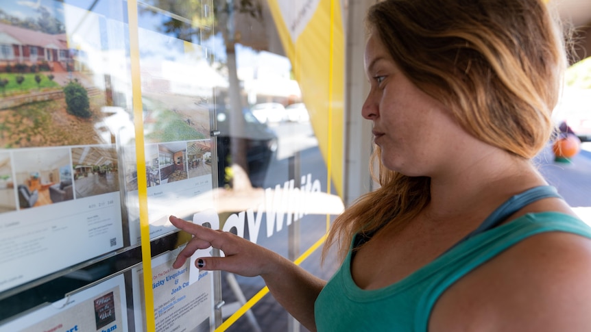 A woman standing side-on points at a window displaying rental listings