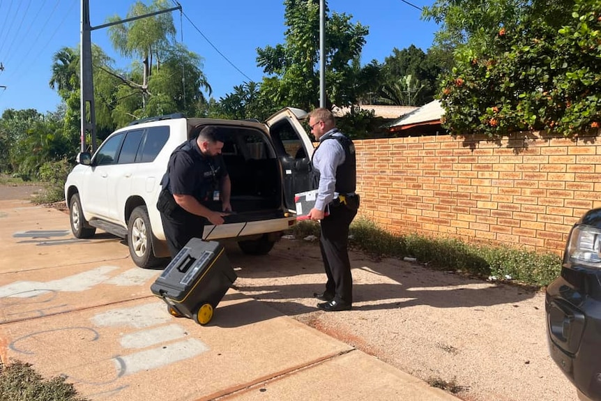Two police unload a large box from the white car outside a house.