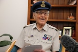 An older man in a military hat smiles at reporters.