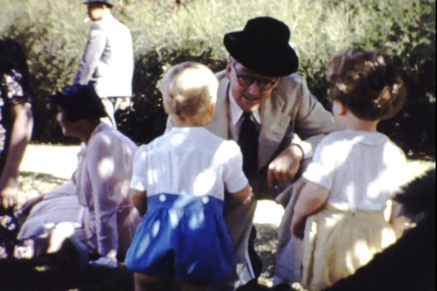 An old grainy photo of a man in a top hat crouched before two toddlers 