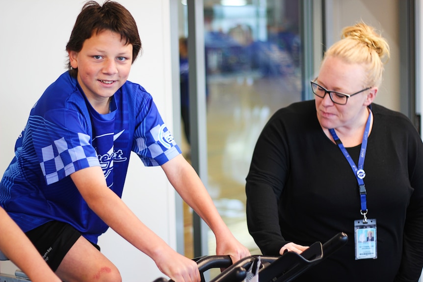 A young boy wearing a blue t-shirt sits on a stationary bike with a woman next to him helping him out.