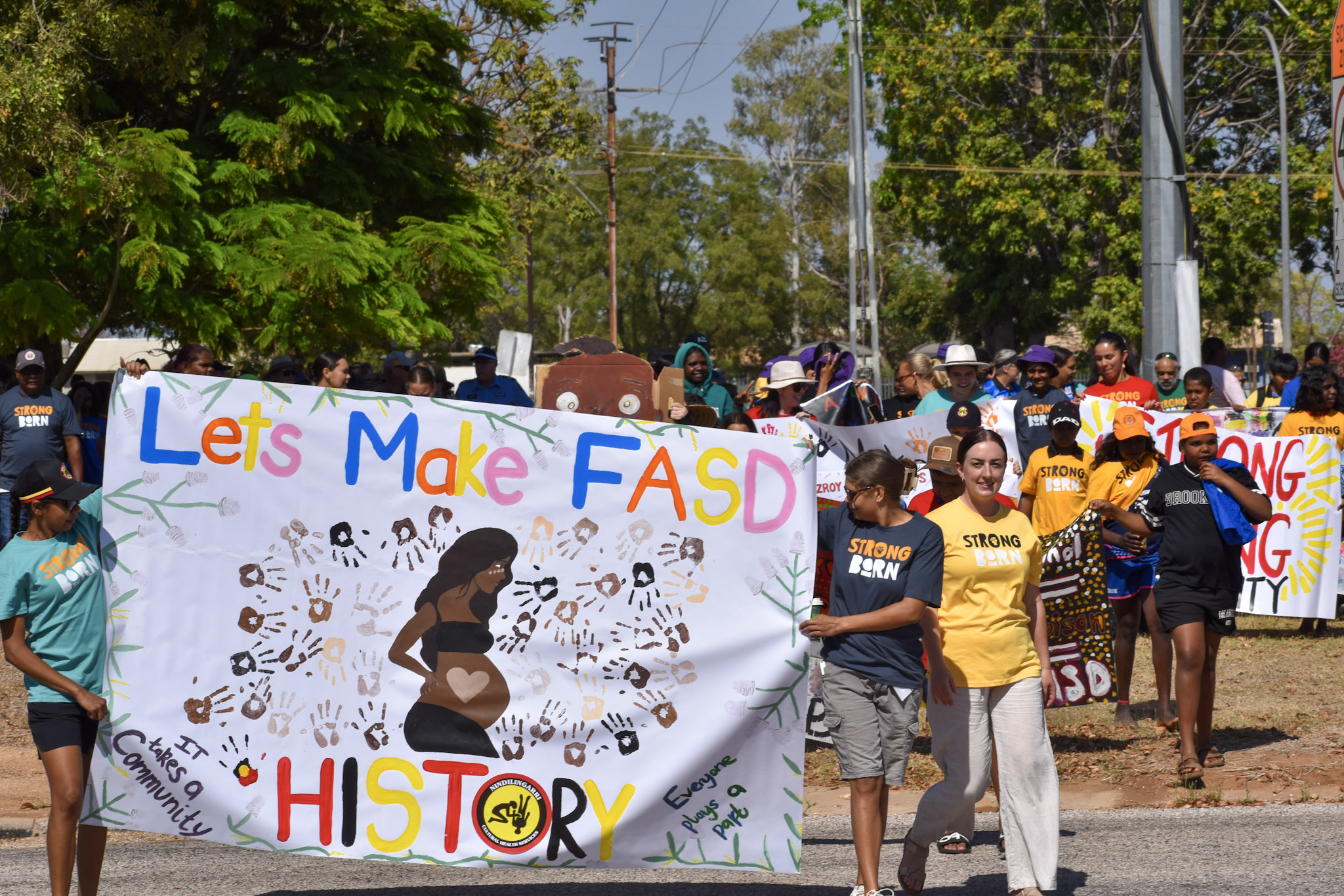 A large, colourful banner with "Let's make FASD History" is carried by Fitzroy Crossing locals, while others march behind.