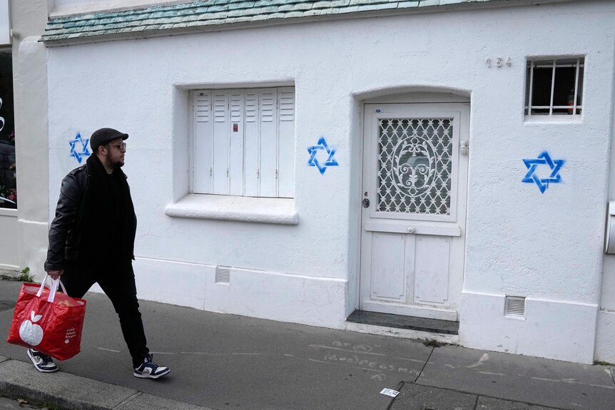 A man walks past a white house with three Stars of David tagged on its wall in blue
