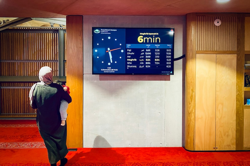 A woman holding a toddler walks past a clock listing the time and prayer times inside a mosque.