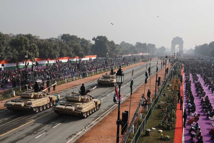 Three tanks drive up a long street. Behind fences, crowds of people watch on. Indian flags are visible