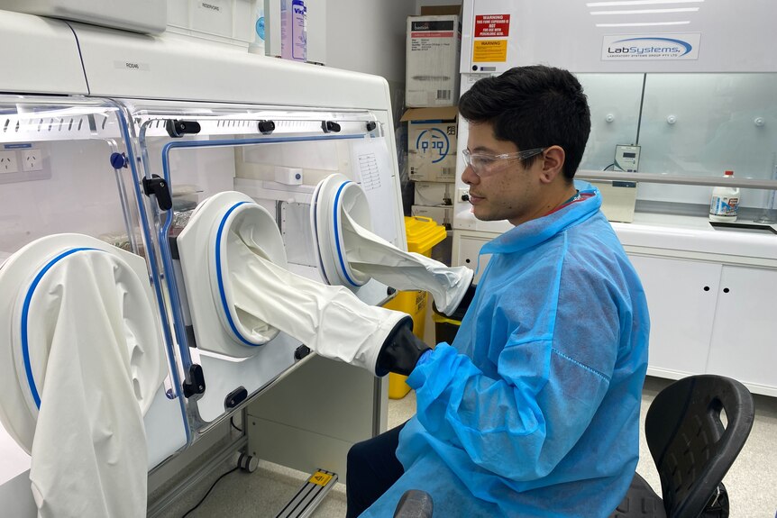 A man in a lab coat puts his hands inside a machine with a glass front