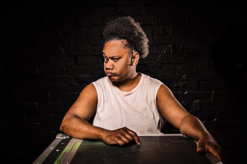 Colour photograph of performer Ghenoa Gela looking unimpressed and standing in front of a dark brick wall.