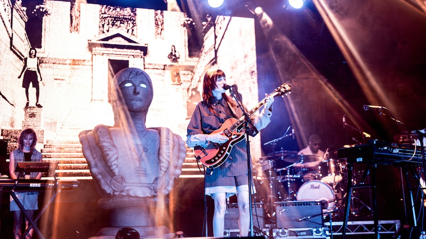 faye stands on stage holding a guitar with other band members and an image of a concrete statue on the screen behind