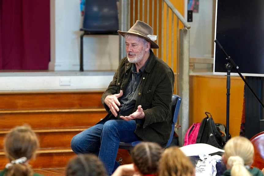 A white mean wearing a hat and brown jacket speaking to a group of kids in a hall. 