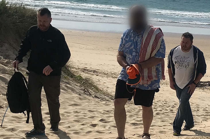 Three men, centre one with blurred face walking away from the beach.