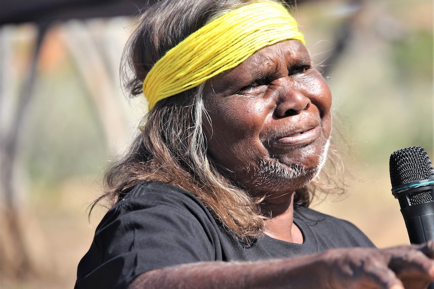 An Indigenous woman with a yellow band around her head look sad while holding a microphone