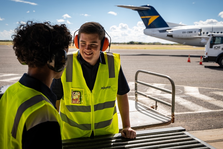 A boy in a high-vis vest smiles while standing on the tarmac. 