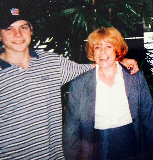 A young man with his arm around his mum. He is wearing a hat a striped polo. She has bright red hair. 