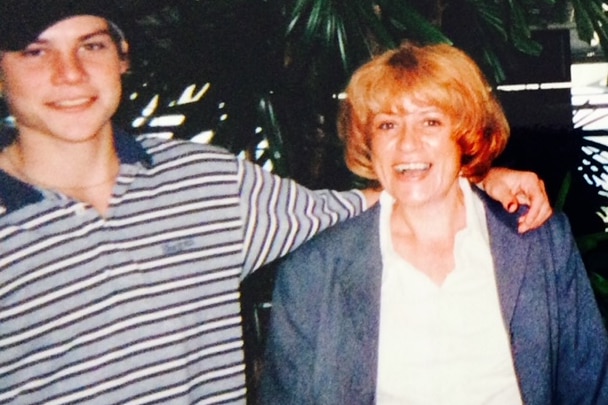 A young man with his arm around his mum. He is wearing a hat a striped polo. She has bright red hair. 