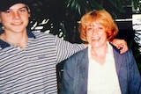 A young man with his arm around his mum. He is wearing a hat a striped polo. She has bright red hair. 