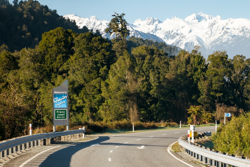 A road with the alps in the background and a 'glacier country' sign visible to the left. 