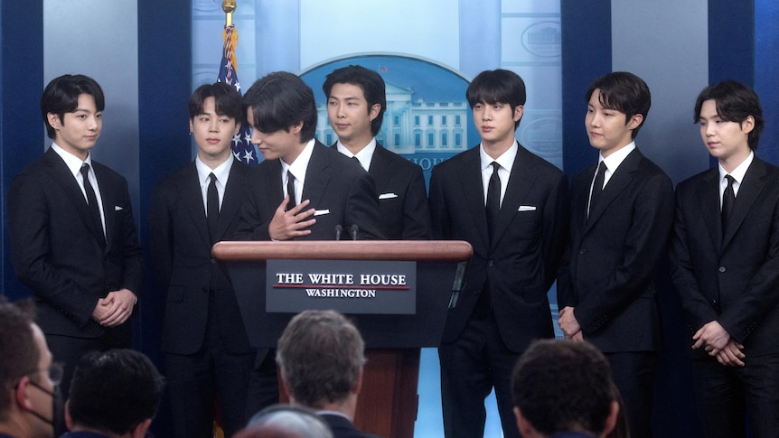 Members of BTS wearing suits stand behind the White House lectern.