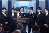 Members of BTS wearing suits stand behind the White House lectern.