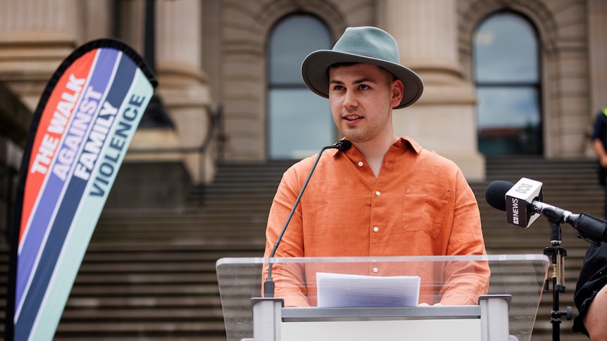 The subject of the story, dark features, wearing a green hat and orange shirt speaking into a microphone in front of Parliament 