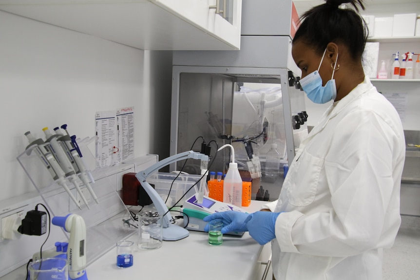 An African woman wearing a lab coat in a lab picks up a small beaker, surrounded by medical equipment
