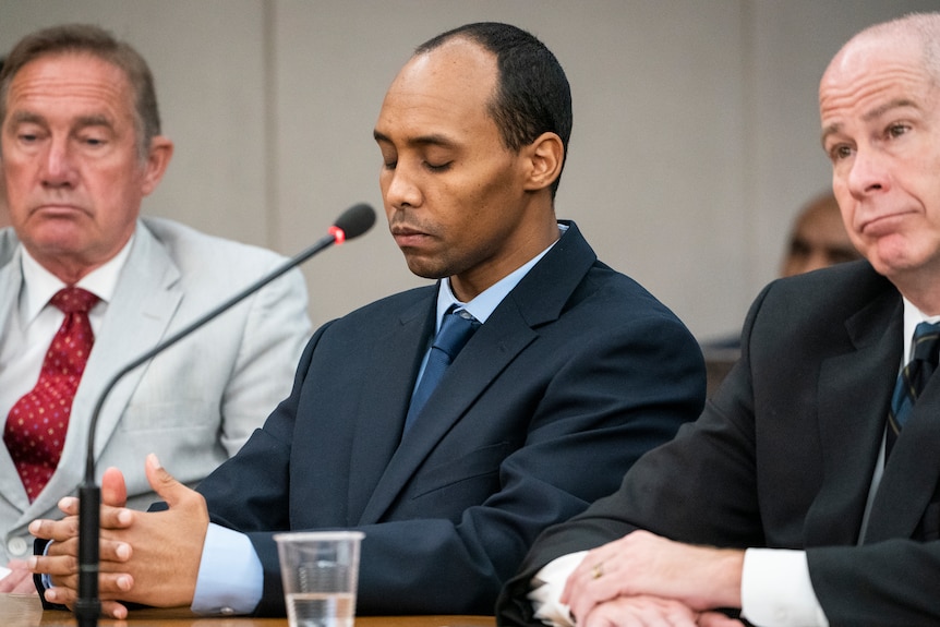 Three men sit at a table in front of a court microphone looking solemn.