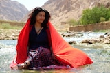 Dark haired woman in colourful flowing clothes sits in front of mountains 