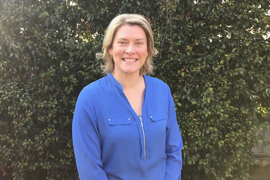 Smiling woman with blonde hair and blue shirt, standing in front of a green hedge.