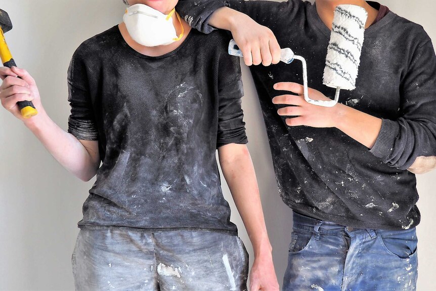 two young people (faces unseen) stand together in paint splattered clothes, holding a mallet and a paint roller
