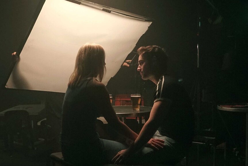 A female and male actress sit in a bar on stools with a light board in front of them while shooting scenes for Exposed