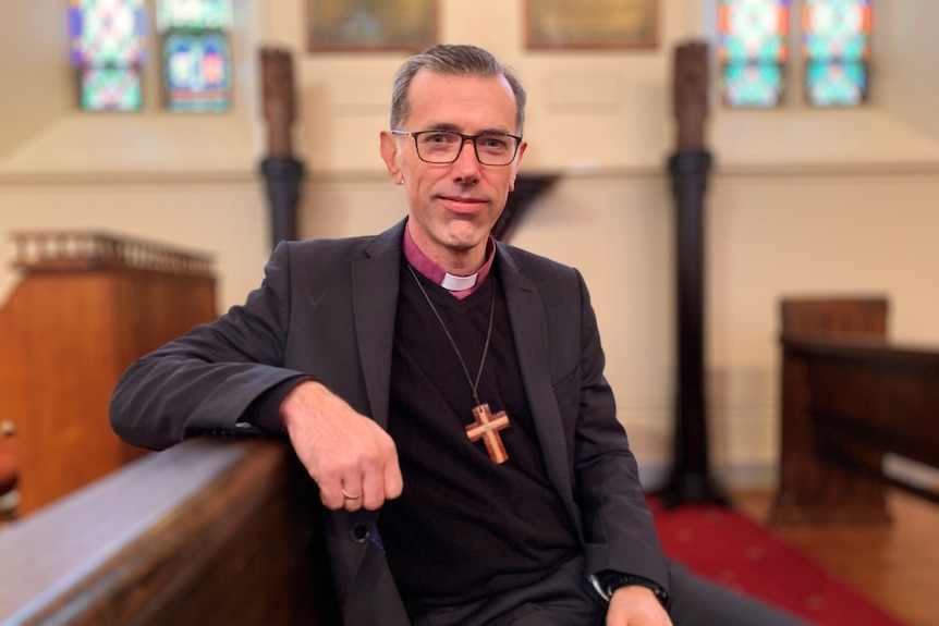 A man wearing a suit jacket and a crucifix on a chain sits on a pew in a church and looks into the camera.