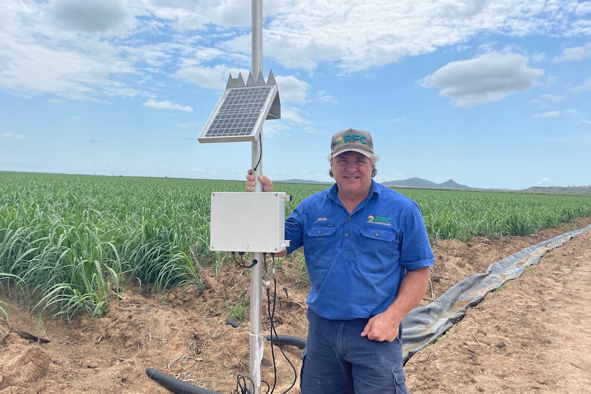 Man in blue workshirt stands in front of cane crop with hand on solar panel device