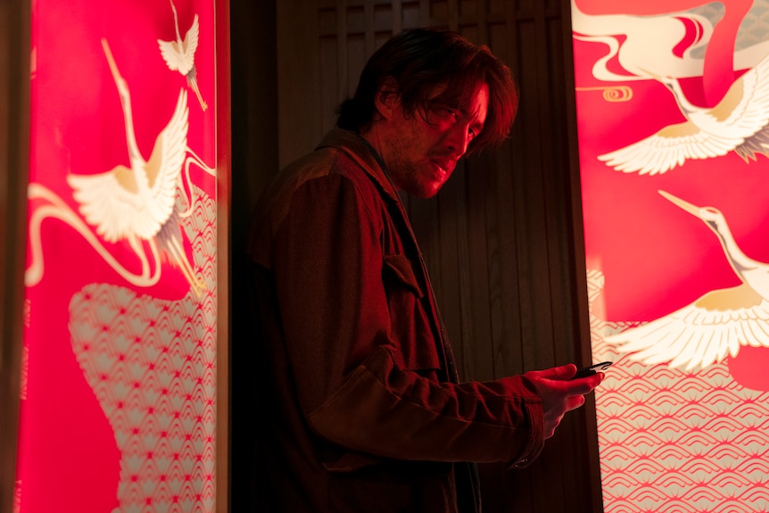 Asian man with dark hair and stubble wears dark jacket and stands in doorway holding phone flanked by bright pink walls.