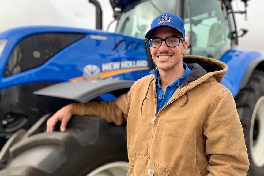 A man in brown jacket and blue cap stands in front of a blue tractor.