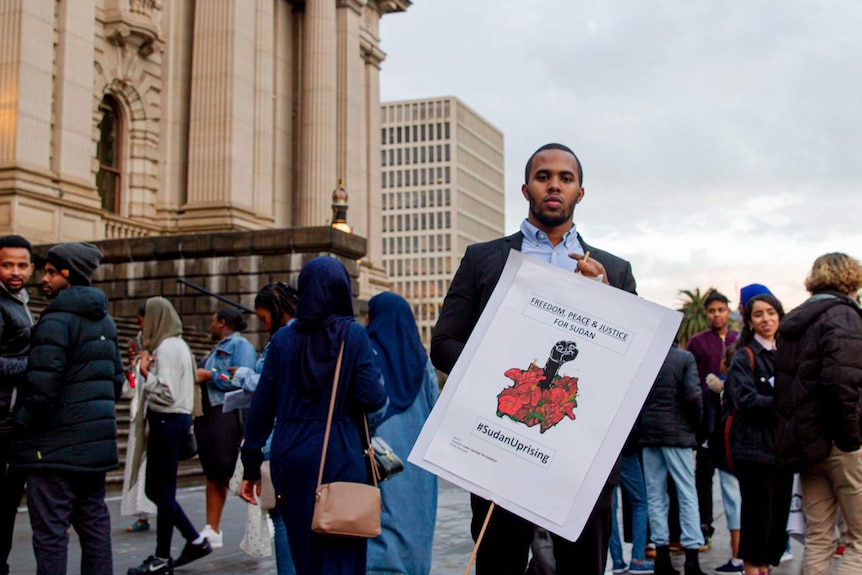 Beside Victoria's neoclassical parliament, a Sudanese-Australian man stands with a 'Sudan Uprising' sign in front of a crowd.